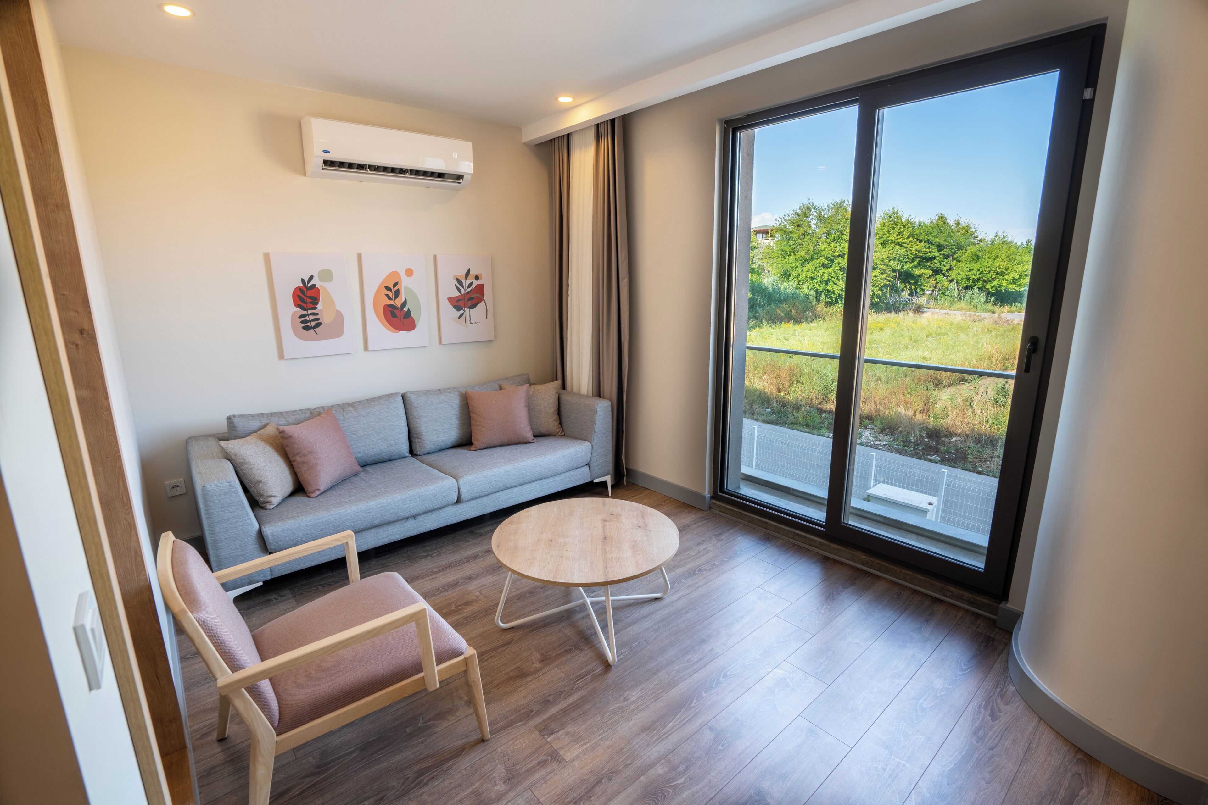 A modern living room with a grey sofa, a round wooden coffee table, and two chairs. There is a large window with a view of greenery outside.