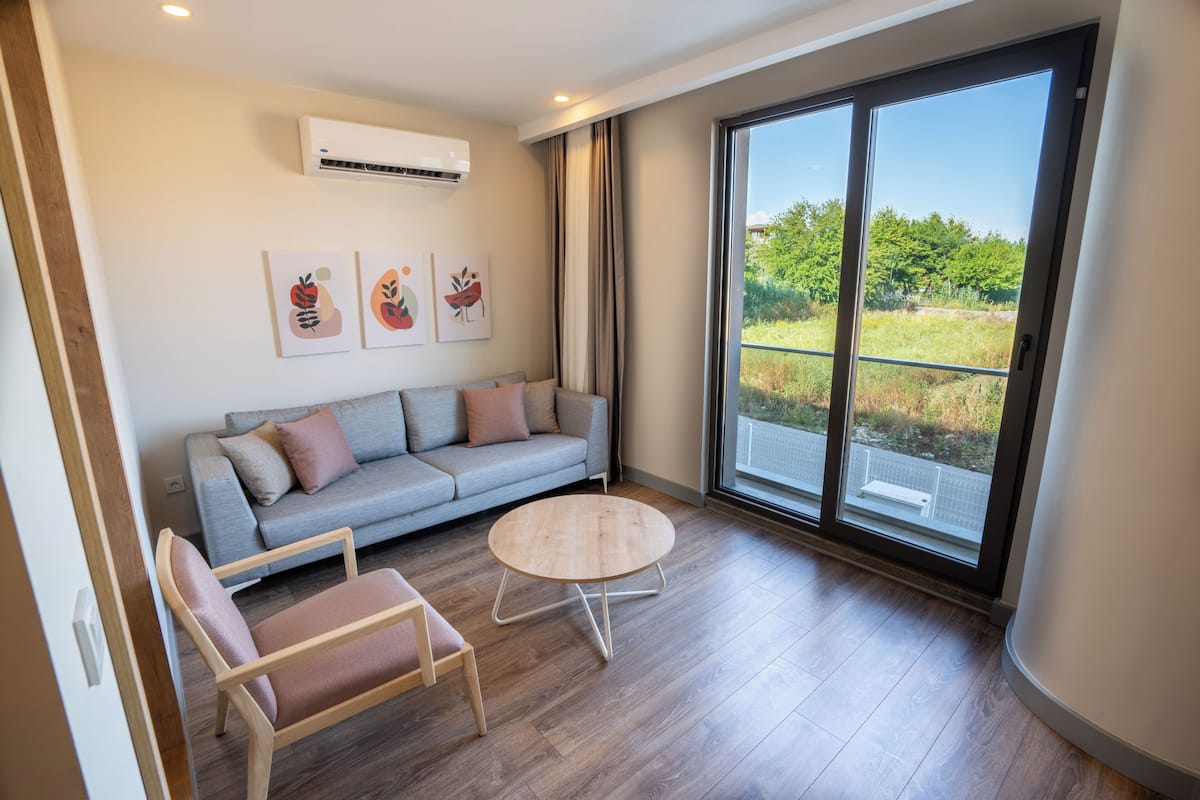 A modern living room with a grey sofa, a round wooden coffee table, and two chairs. There is a large window with a view of greenery outside.
