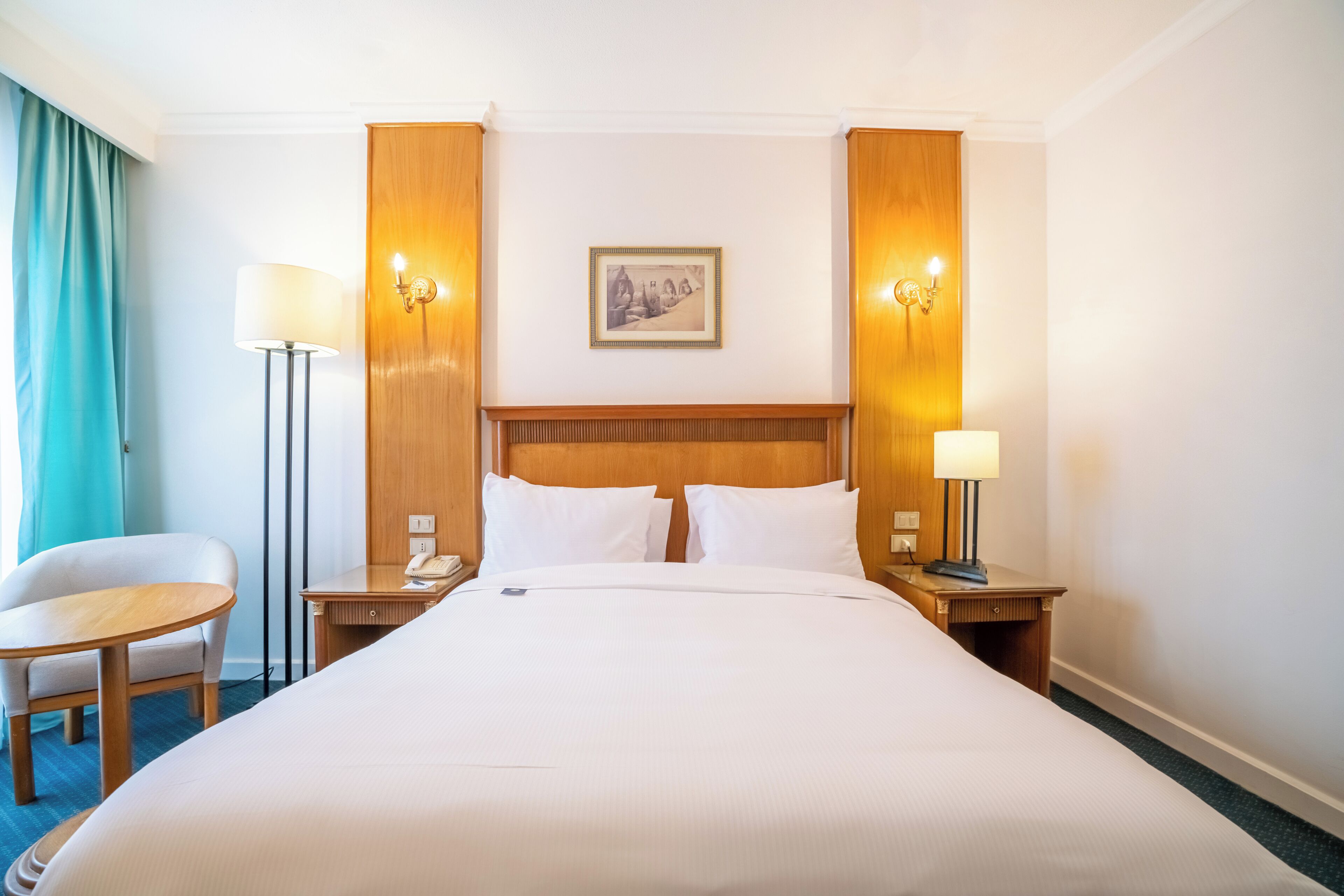 A hotel room with a large bed, two bedside tables with lamps, a small round table with a chair, and a framed picture on the wall.