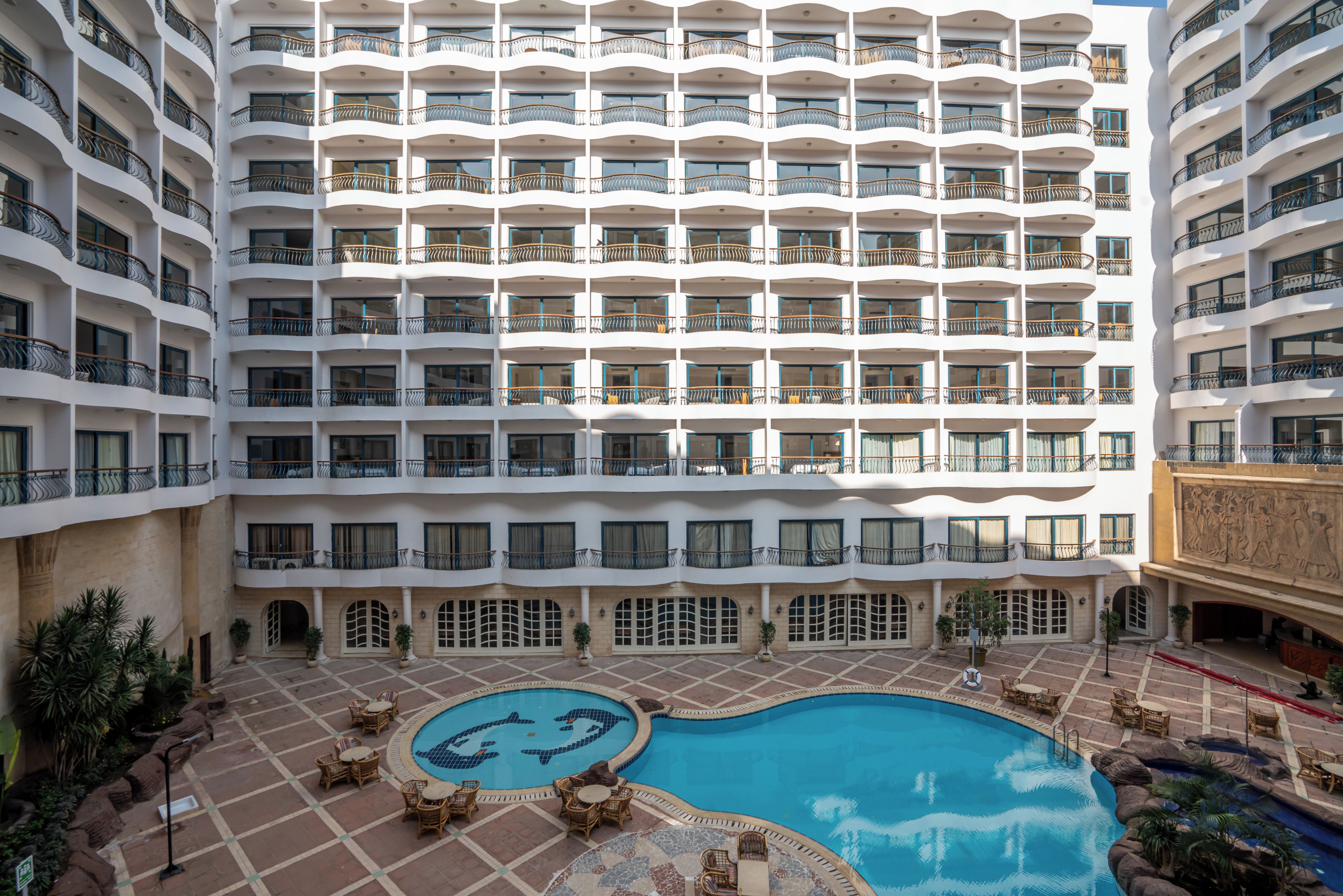 Ein mehrstöckiges Hotel mit einem zentralen Schwimmbecken, umgeben von Sitzbereichen im Freien und Kübelpflanzen.