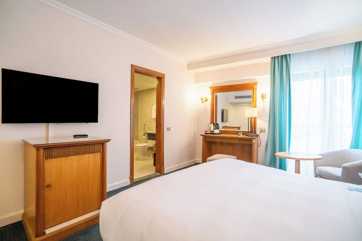 A hotel room with a large bed, a TV mounted on the wall, a wooden wardrobe, a bathroom visible through an open door, a small round table, and a chair.