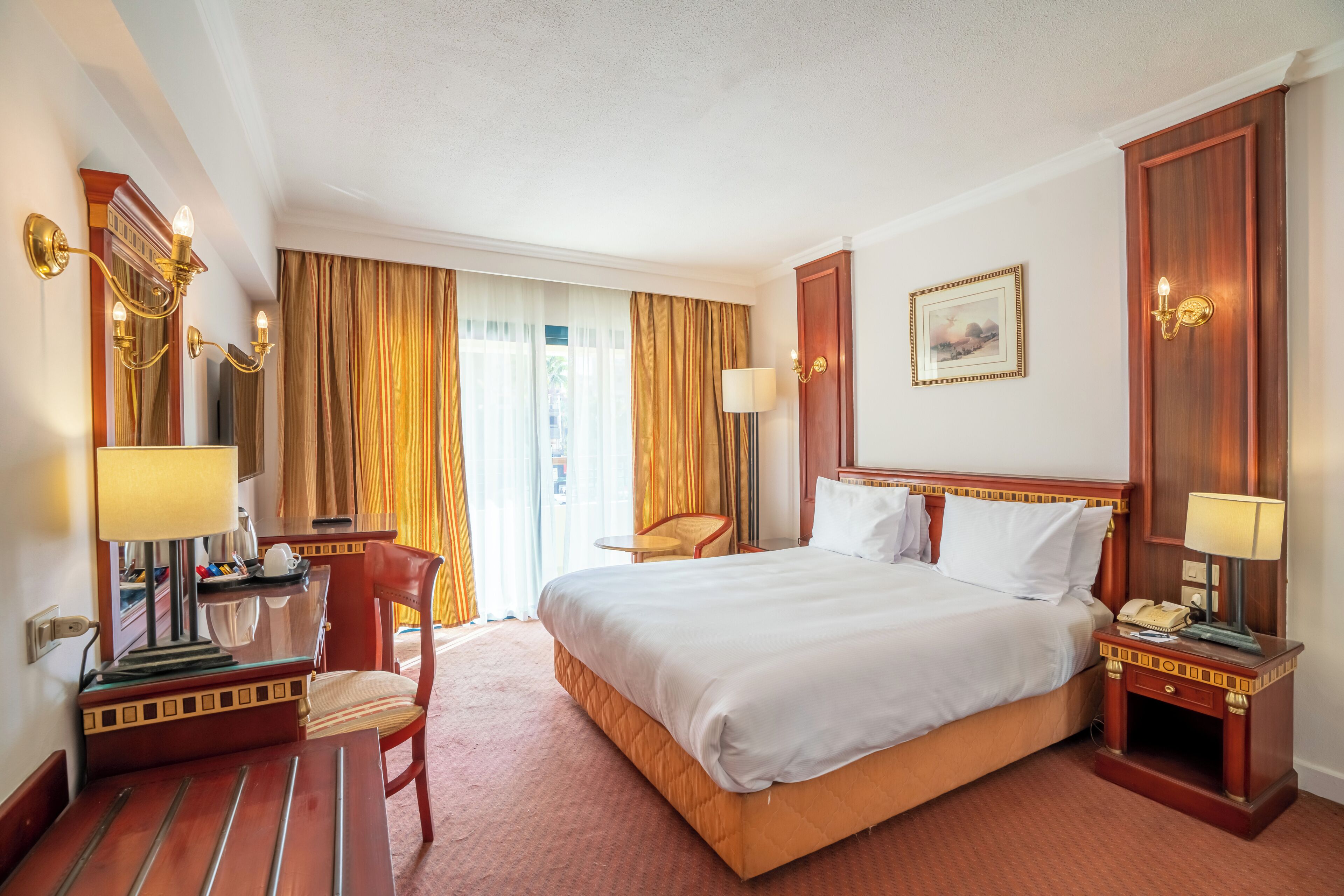 A hotel room with a large bed, a desk with a lamp, a chair, and a nightstand with a phone.