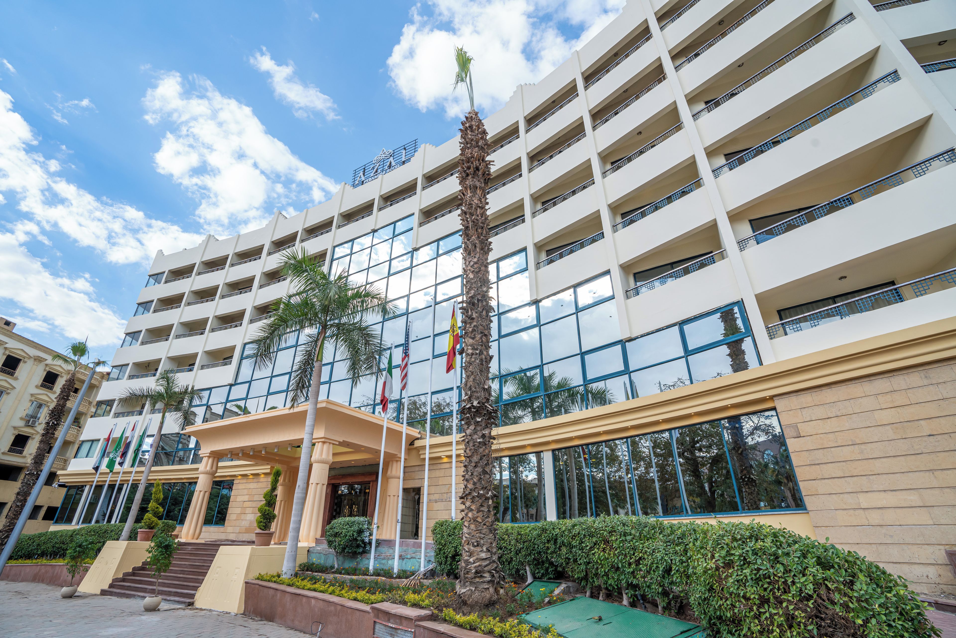Un hotel de varios pisos con un diseño moderno, que presenta una entrada central con escalones, palmeras y un jardín bien cuidado.