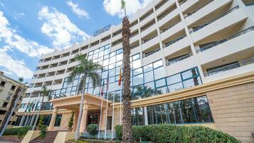 Un hotel de varios pisos con un diseño moderno, que presenta una entrada central con escalones, palmeras y un jardín bien cuidado.