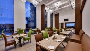 A modern dining area with multiple tables and chairs, large windows, and a flat-screen TV mounted on the wall.