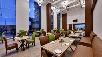 A modern dining area with multiple tables and chairs, large windows, and a flat-screen TV mounted on the wall.