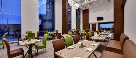 A modern dining area with multiple tables and chairs, large windows, and a flat-screen TV mounted on the wall.