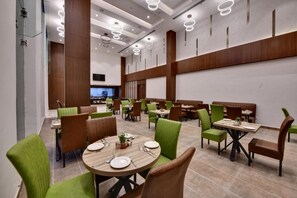 A modern restaurant with round tables, green and brown chairs, and a large TV screen.