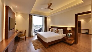 A modern hotel room with a large bed, a desk, a chair, a TV, a ceiling fan, and a view of the city.