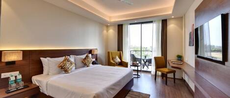 A modern hotel room with a large bed, a desk, a chair, a TV, and a balcony with a view.