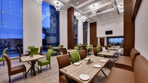 A modern dining area with multiple tables and chairs, large windows, and a flat-screen TV mounted on the wall.