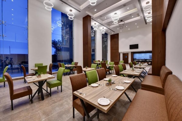 A modern dining area with multiple tables and chairs, large windows, and a flat-screen TV mounted on the wall.
