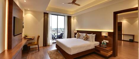 A modern hotel room with a large bed, a desk, a chair, a TV, a ceiling fan, and a view of the city.