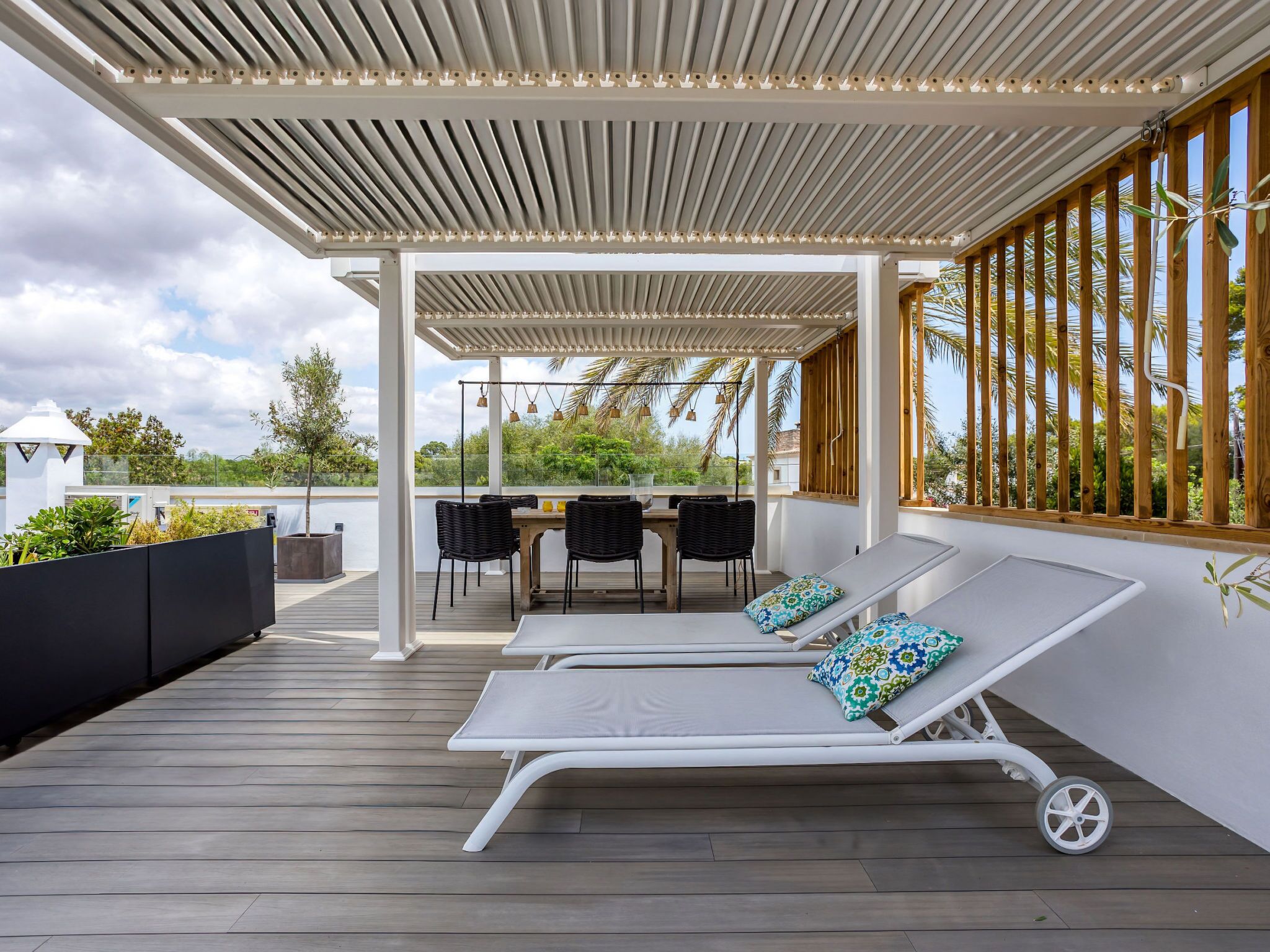 A covered outdoor seating area with lounge chairs and a table, surrounded by wooden columns and a view of greenery.