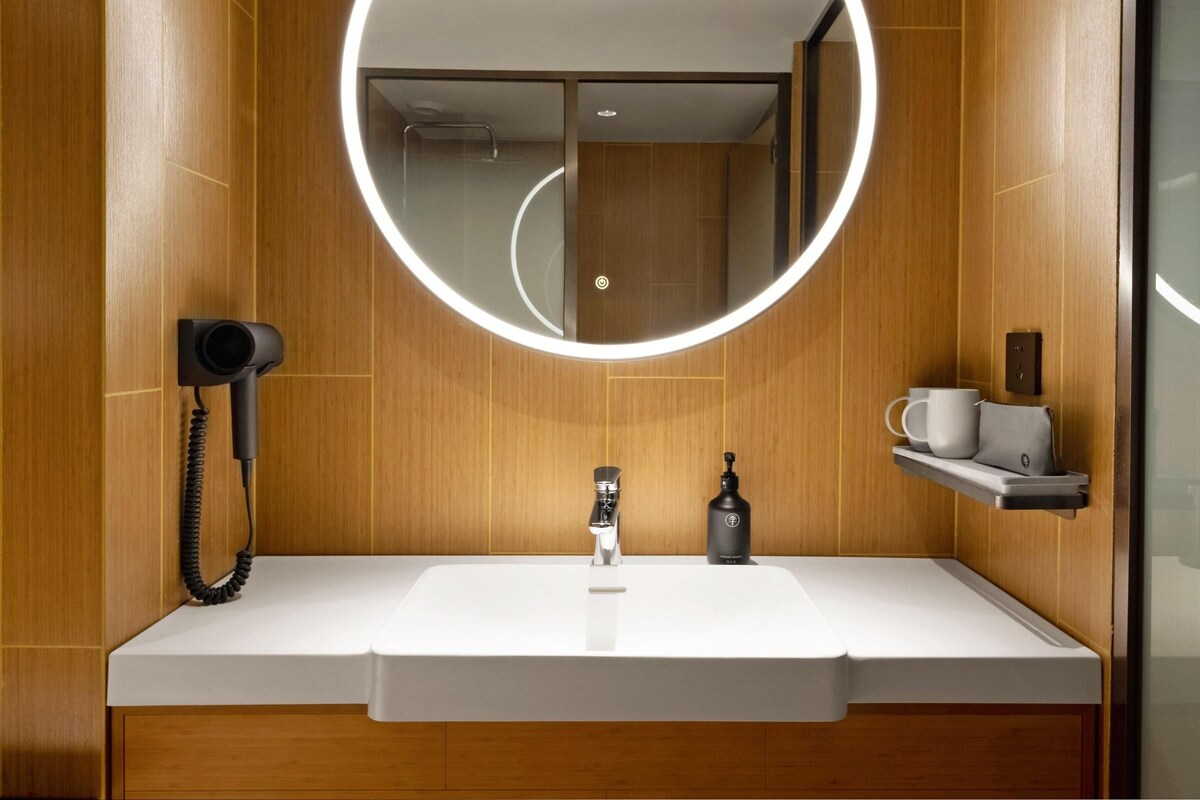 Ein modernes Badezimmer mit einem weißen Waschbecken, einem runden Spiegel mit Beleuchtung, einem Fön und einer kleinen Ablage mit Tasse und Flasche.