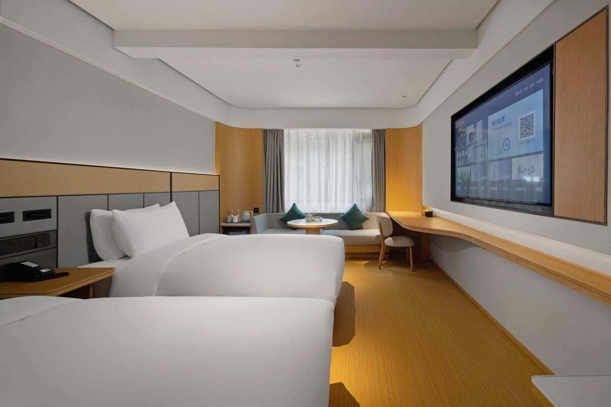 A hotel room with a bed, a desk, a chair, a small table, and a TV mounted on the wall.