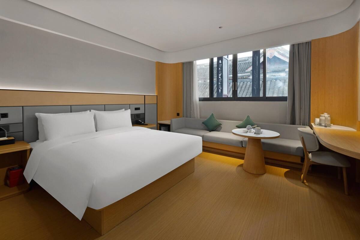 A modern hotel room with a large bed, a sofa, a small dining table, and a view of traditional architecture through the window.