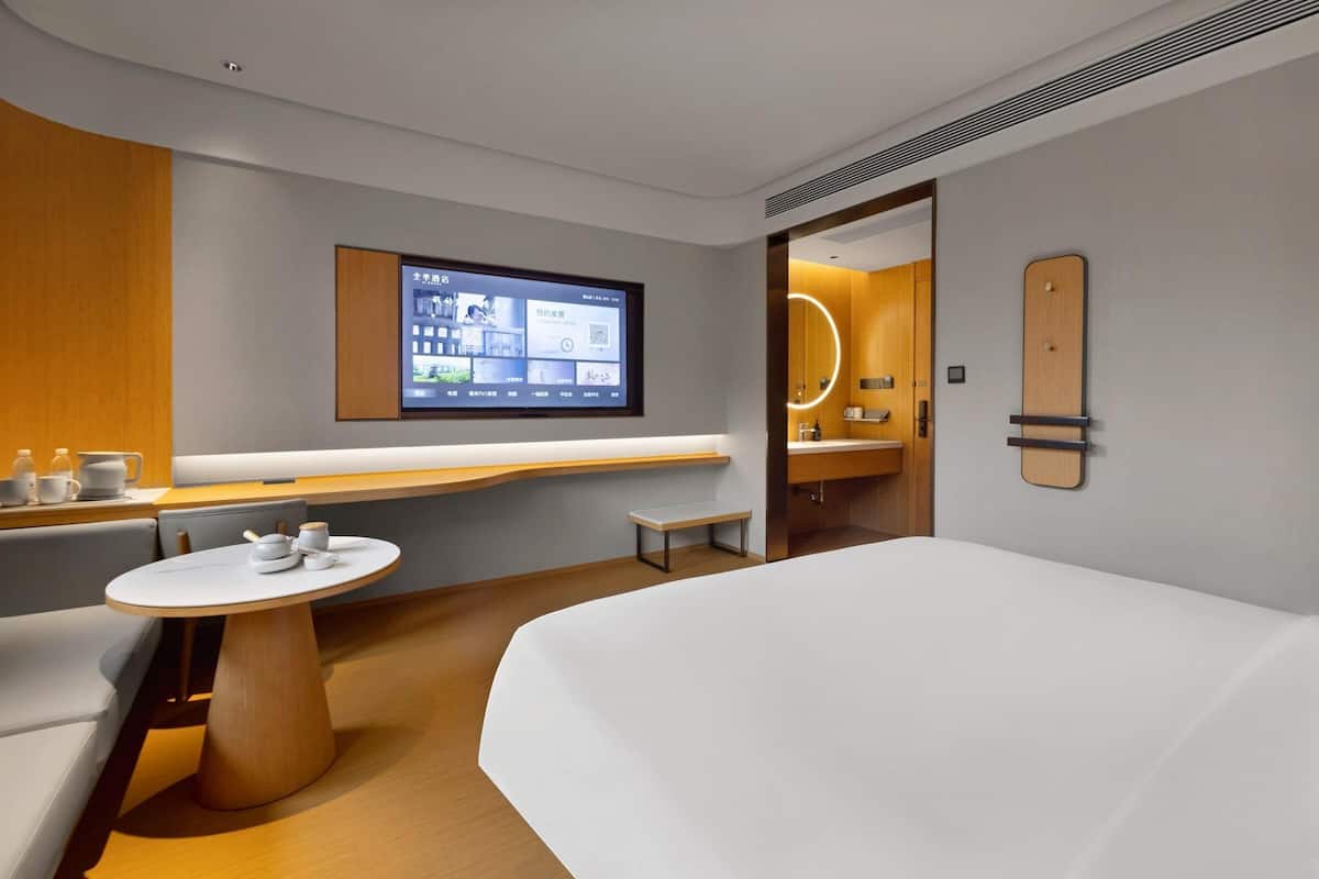 A modern hotel room with a bed, a round table with a white tablecloth, a built-in TV, and a wooden accent wall.