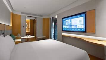 A modern hotel room with a large bed, a desk, a chair, a small table with a cup, a mirror, and a TV displaying a QR code.