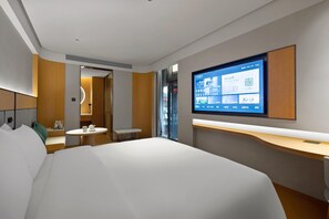 Ein modernes Hotelzimmer mit einem großen Bett, einem Schreibtisch, einem Sessel, einem kleinen Tisch mit einer Tasse, einem Spiegel und einem Fernseher, der einen QR-Code anzeigt.