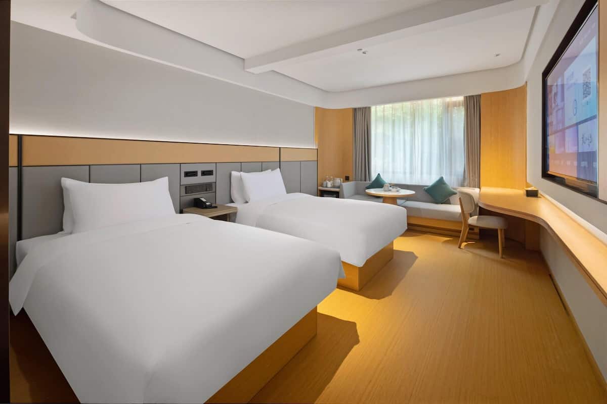 Quarto de hotel com duas camas, uma mesa, uma cadeira, uma televisão e uma janela com cortinas.