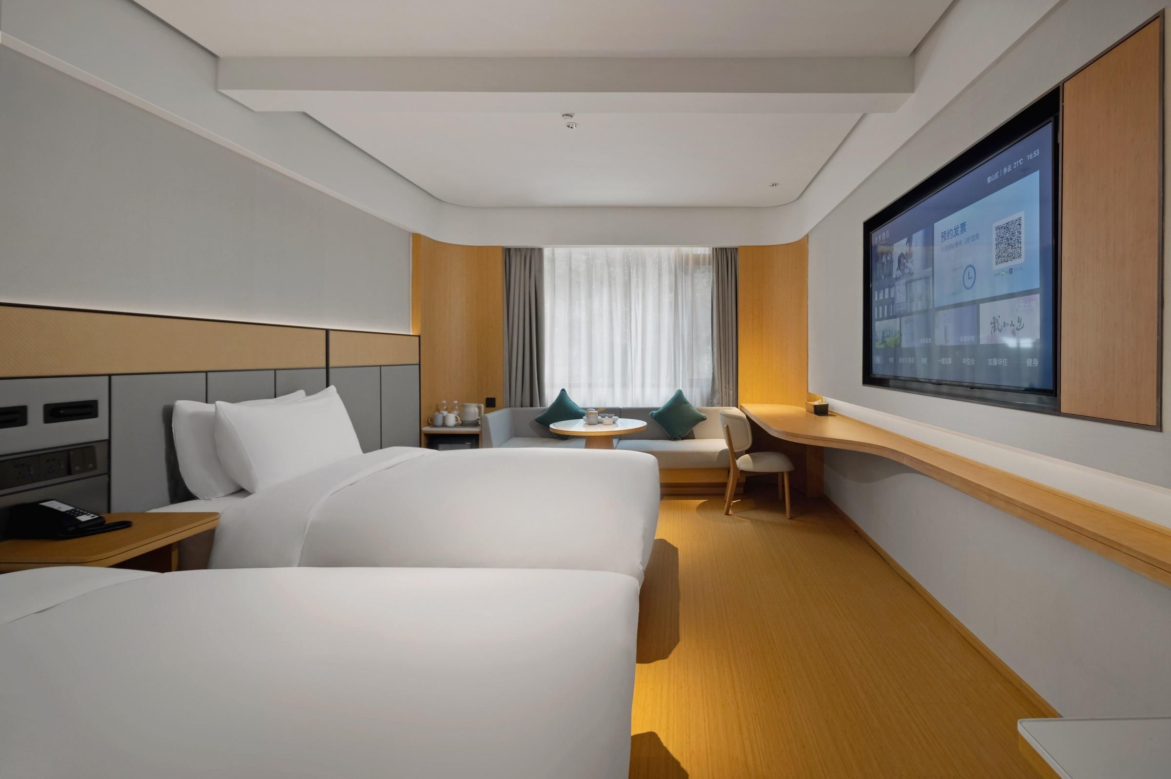 A hotel room with a bed, a desk, a chair, a small table, and a TV mounted on the wall.