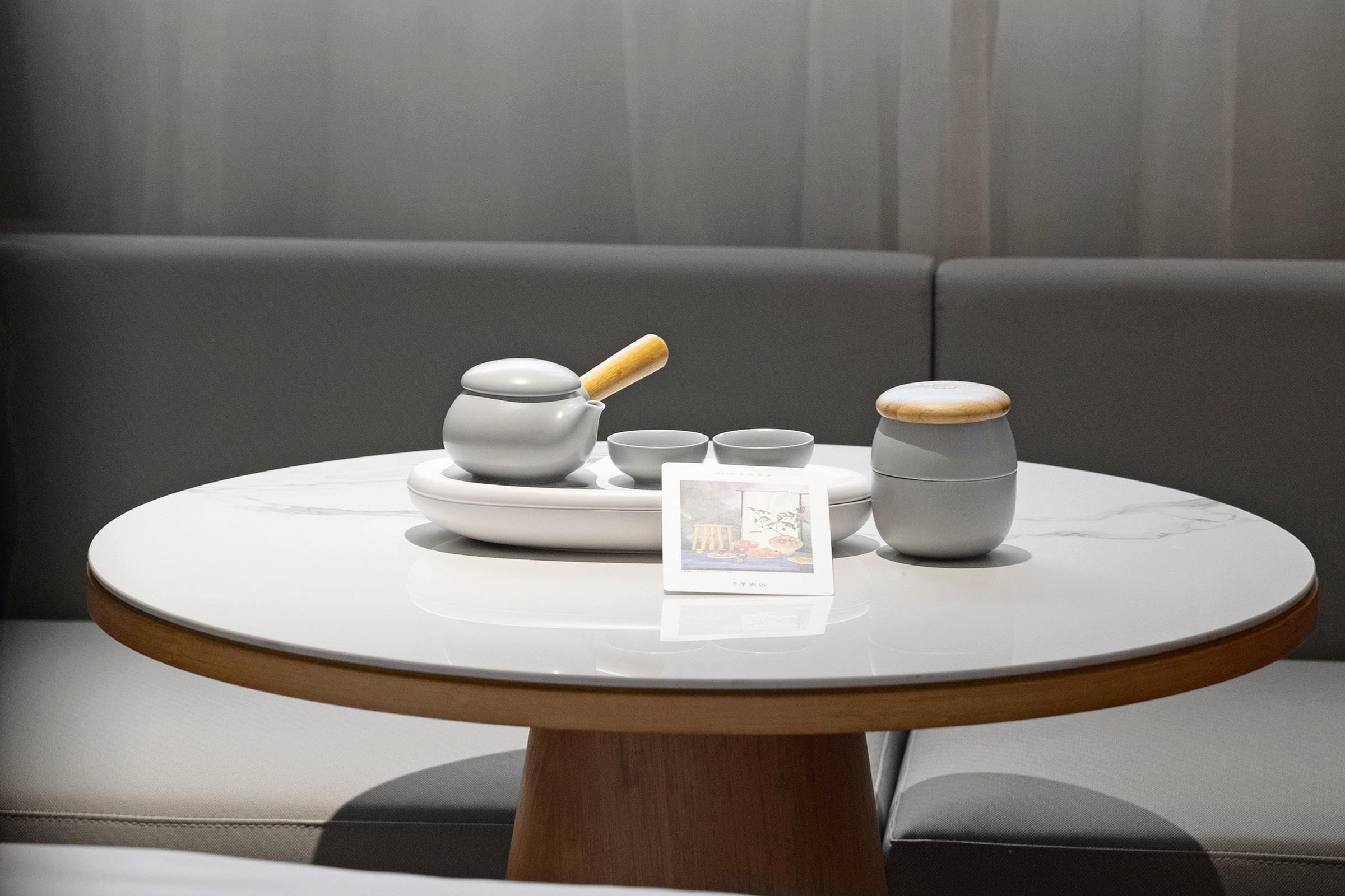 A round table with a wooden base, a white ceramic pitcher, a small bowl, and a cup, set against a grey sofa backdrop.