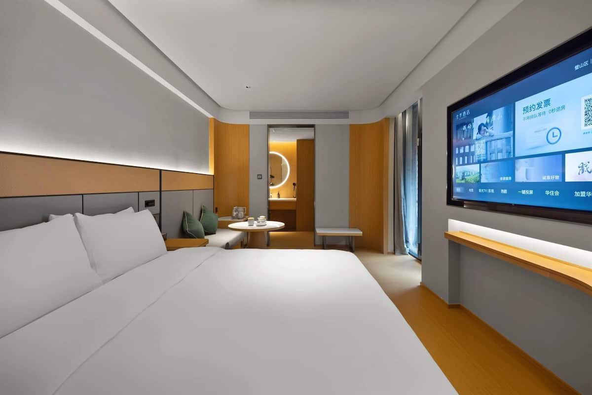 A modern hotel room with a large bed, a TV, a small table, and a seating area.