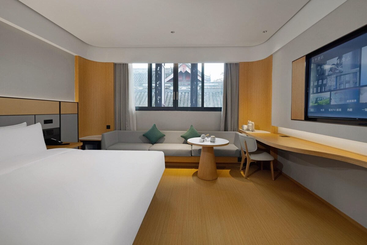 Ein Hotelzimmer mit Bett, Sofa, Schreibtisch, Fernseher und einem Fenster mit Blick auf Gebäude.