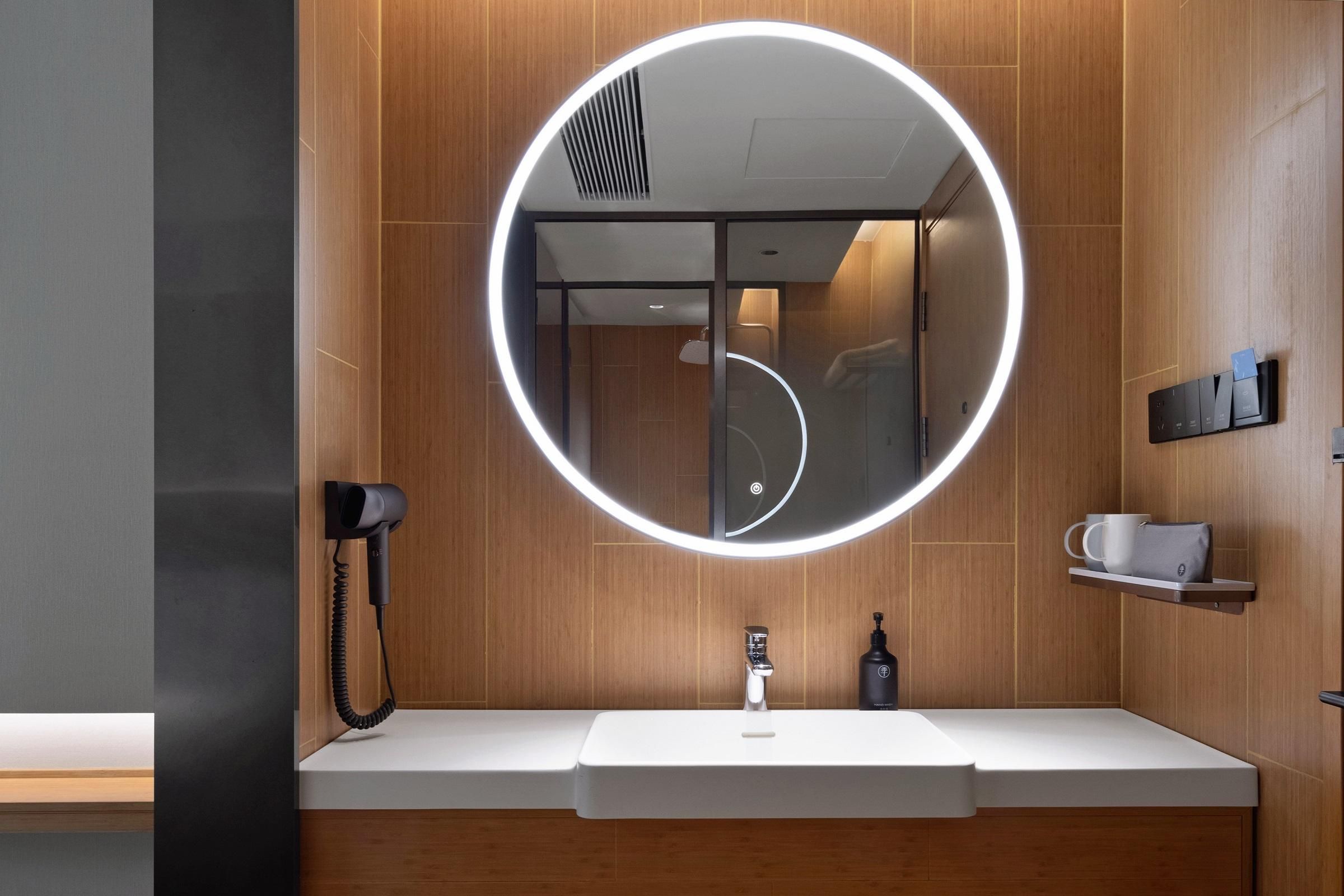 A modern bathroom with a round mirror, a hairdryer, a sink, and a small shelf with a cup and a bottle.
