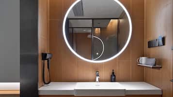 A modern bathroom with a round mirror, a hairdryer, a sink, and a small shelf with a cup and a bottle.