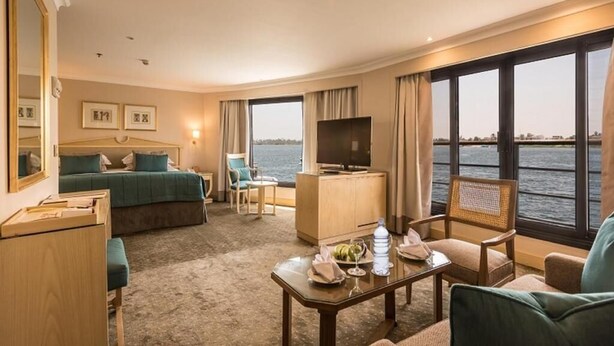 A hotel room with a bed, a TV, a desk, a chair, and a view of the water. - Steigenberger Regency Cruise 4&7 Nights From Luxor (Luxor)