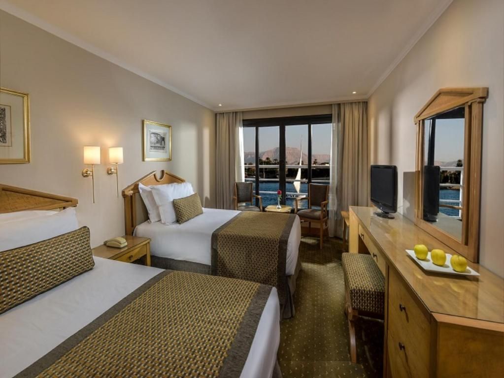 Quarto de hotel com duas camas, uma escrivaninha, uma cadeira, uma televisão e uma grande janela vista para o mar.