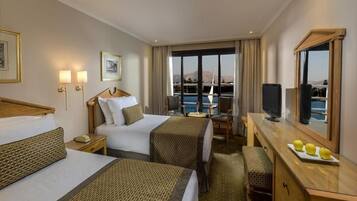 A hotel room with two beds, a desk, a chair, a TV, and a large window with a view of the sea.