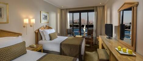Quarto de hotel com duas camas, uma escrivaninha, uma cadeira, uma televisão e uma grande janela vista para o mar.