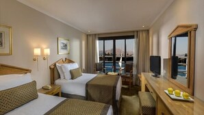 A hotel room with two beds, a desk, a chair, a TV, and a large window with a view of the sea. - Steigenberger Regency Cruise 4&7 Nights From Luxor (Luxor)