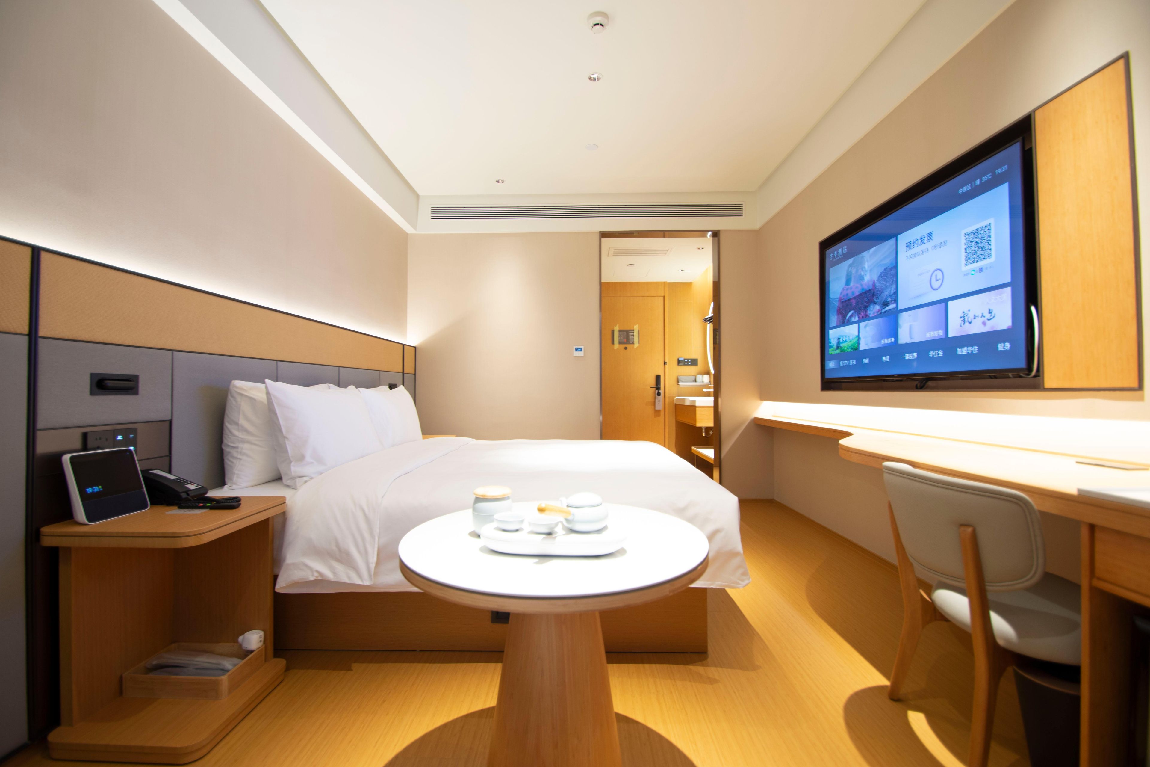 A hotel room with a bed, bedside table with a phone, a desk with a chair, a TV on the wall, and a small table with cups and a tray.