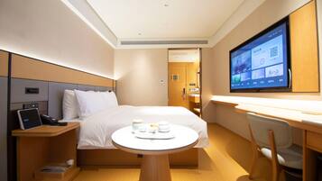 A hotel room with a bed, bedside table with a phone, a desk with a chair, a TV on the wall, and a small table with cups and a tray.