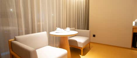 A modern hotel room with a round wooden table, two cushioned chairs, and a large window with sheer curtains.