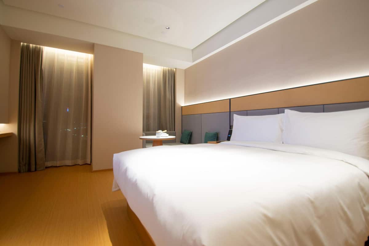 Ein Hotelzimmer mit einem großen Bett, einem Nachttisch, einem Sessel und einem Fenster mit Vorhängen.