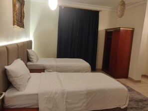 A hotel room with two beds, a nightstand, a wooden wardrobe, and a dark curtain. - Golden Dana Agadir (Jizan)