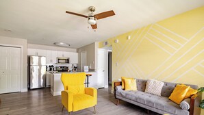 Eine moderne Küche mit weißen Schränken und Edelstahlgeräten, ein Essbereich mit einem gelben Stuhl und einem grauen Sofa mit gelben Kissen sowie ein Deckenventilator.