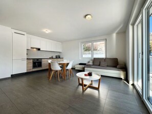 Et moderne køkken med hvide skabe, et spiseområde med et rundt bord og stole, samt en opholdsstue med sofa og et stort vindue.