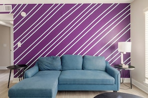 Un salon moderne avec un canapé d’angle, une petite table ronde, une lampe et un mur orné de rayures diagonales.