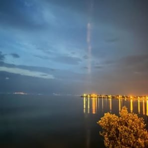 Vista al agua con un muelle que se adentra en aguas tranquilas, un árbol en primer plano y un cielo despejado.