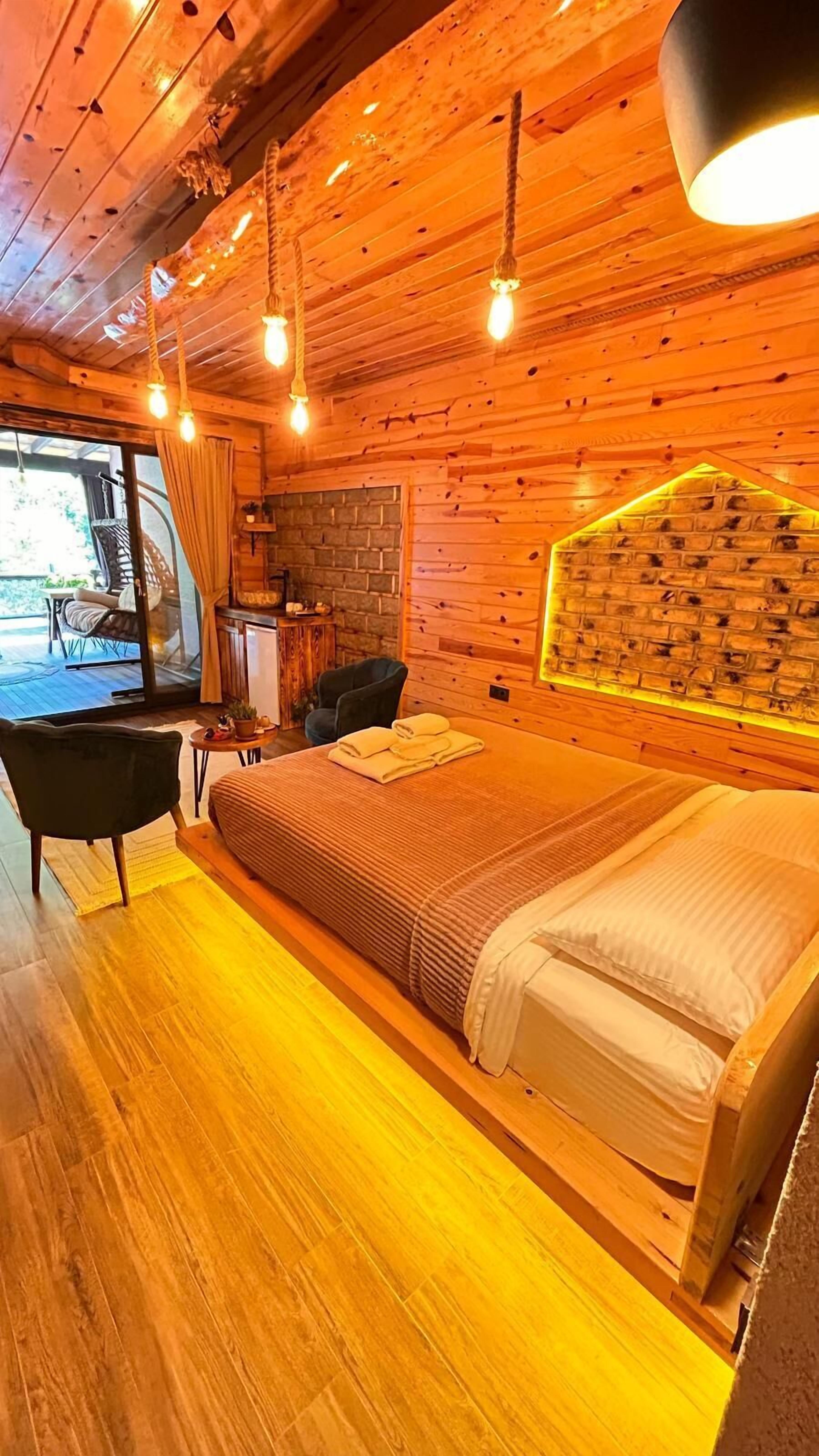 A cozy wooden room with a bed, a chair, a small table, and a view of the outdoors.