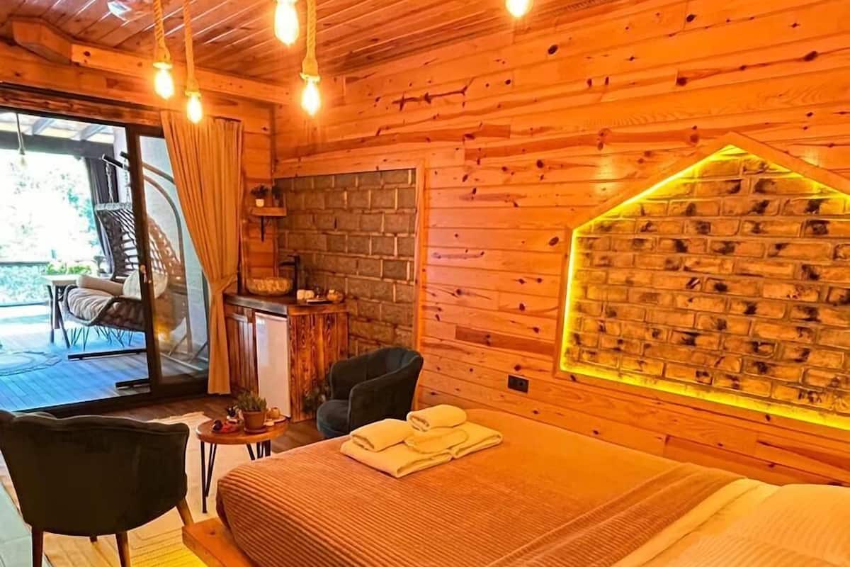 A cozy wooden room with a bed, a chair, a small table, and a view of the outdoors.