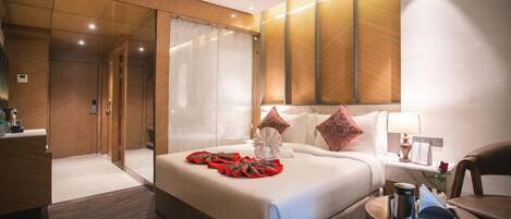 A modern hotel room with a large bed, a bedside table with a lamp, a chair, and a small table with a tray.