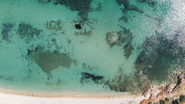 Luftaufnahme eines Strandes mit klarem türkisblauem Wasser, einem Sandstrand und Reihen von Sonnenschirmen.