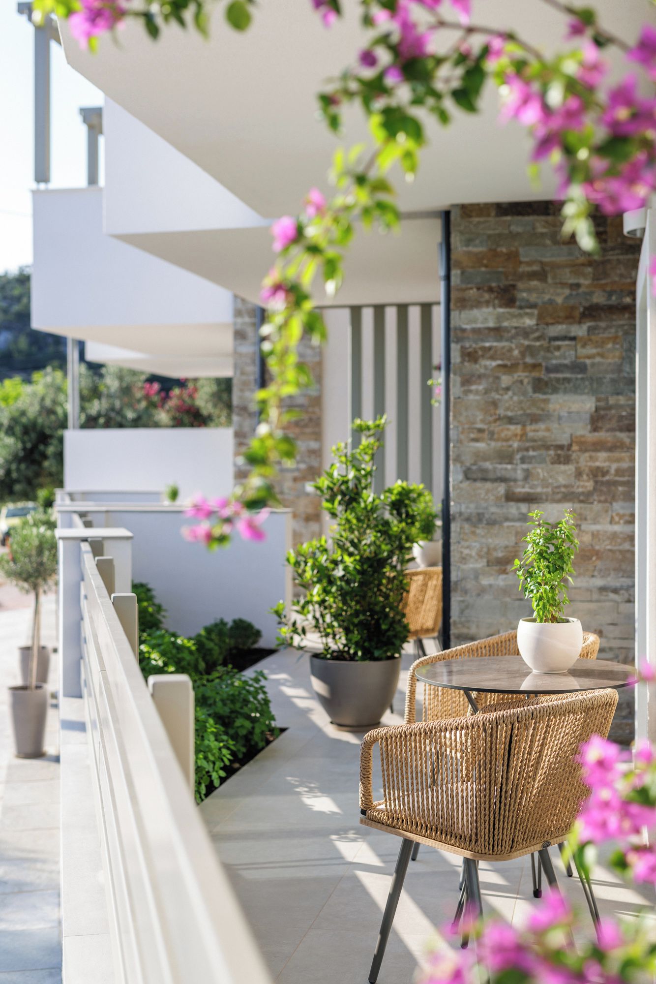 A modern outdoor seating area with wicker chairs and a table, surrounded by potted plants and a stone wall.